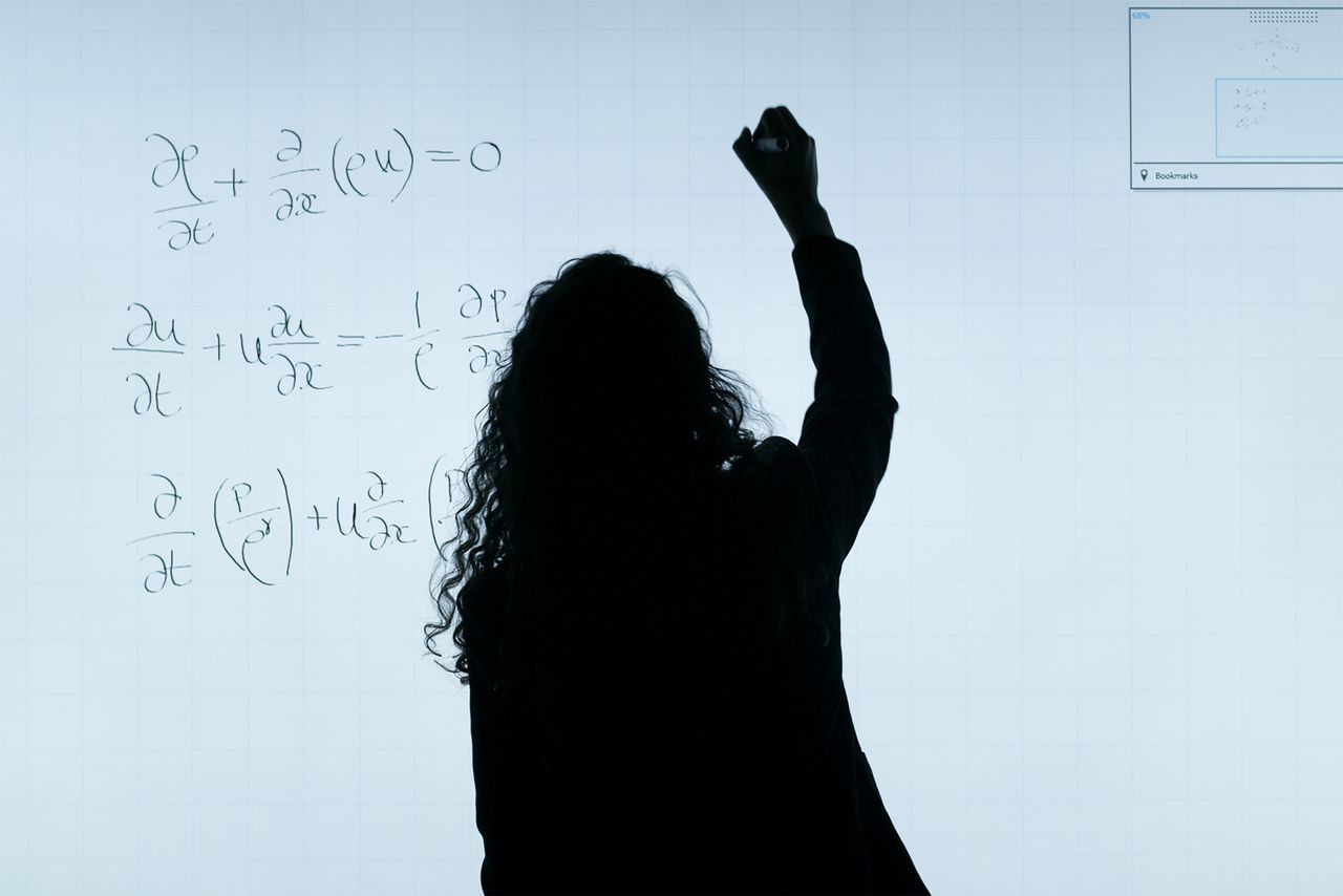 Home A silhouette of a woman writing mathematical equations on a large whiteboard, representing education and analysis.