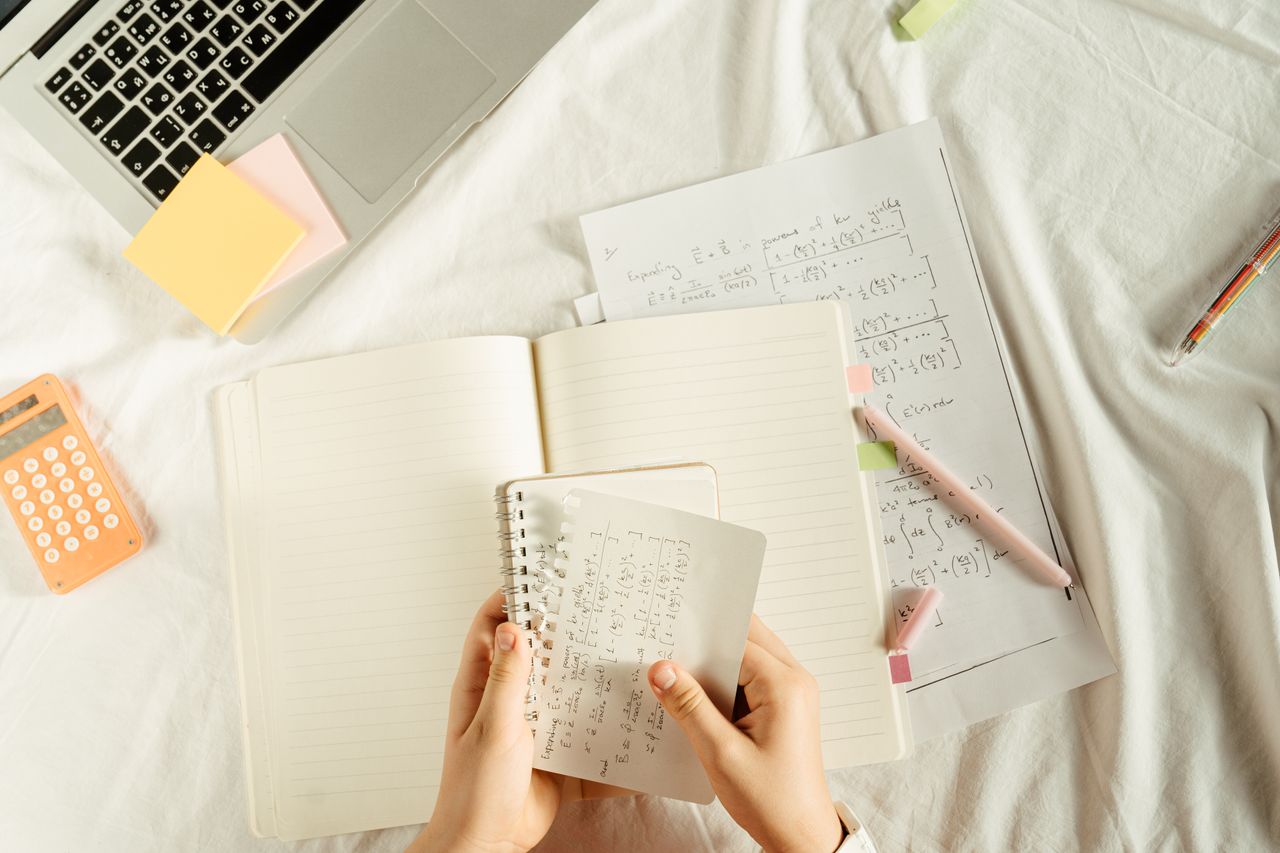 Home Hands holding a notebook with math equations, calculator, and laptop on a study desk.